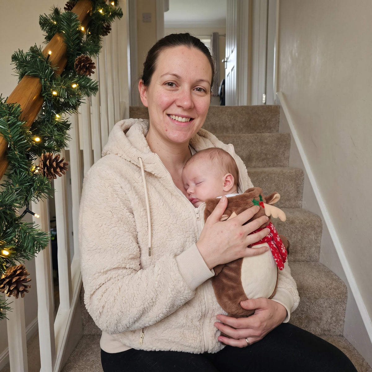 Mum on the stairs at Christmas wearing the Mistletot Reindeer baby pouch hoodie with her baby asleep chest to chest